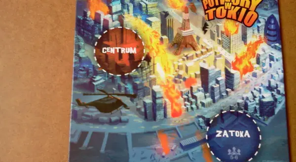 Ilustracja gry planszowej "Potwory w Tokio" z płonącym miastem, wieżą Eiffla i helikopterem.