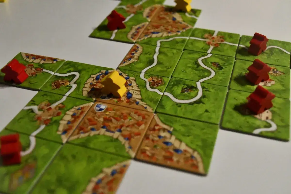 Rozgrywka w Carcassonne. Drewniane pionki w kolorach czerwonym i żółtym stoją na planszy z kafelków przedstawiających krajobraz z drogami i zamkami.