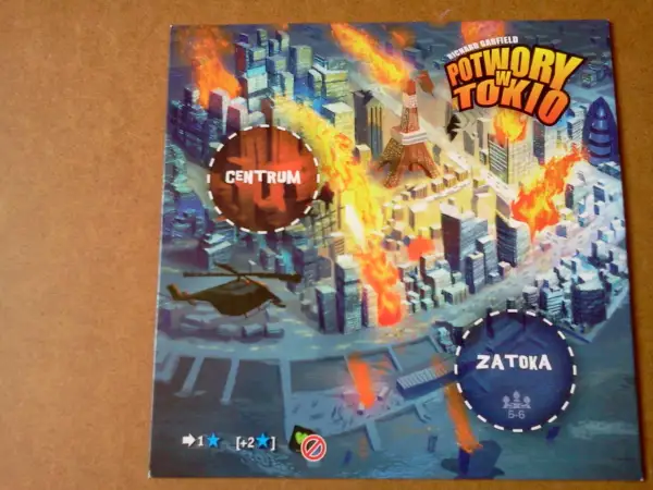 Ilustracja gry planszowej "Potwory w Tokio" z płonącym miastem, wieżą Eiffla i helikopterem.